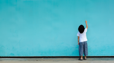 Child standing against a bright blue wall, pointing upward with curiosity.